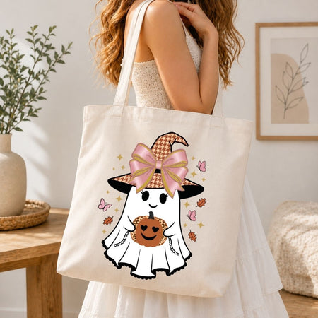A woman wearing a white dress carries a cream canvas tote bag featuring a cute cartoon ghost in a pink bow witch hat holding a pumpkin. The scene is set in a warm, softly lit room with a wooden table, potted plant, and framed wall art in the background.