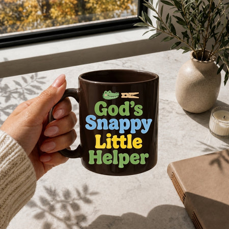 Person holding a mug with 'God's Snappy Little Helper' text on a table with a window and plant in the background