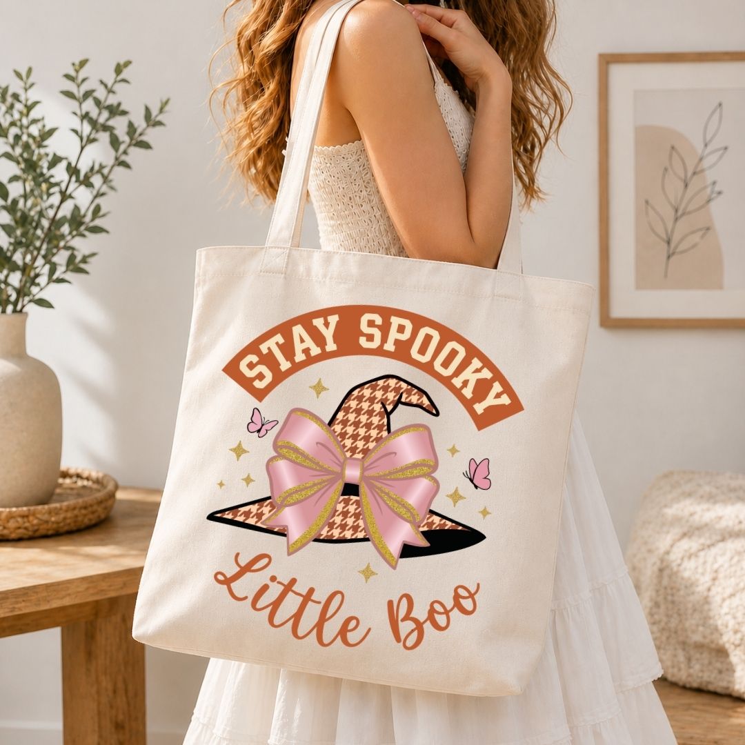A woman in a white dress carries a cream canvas tote bag with a 'Stay Spooky Little Boo' graphic featuring a witch hat and pink bow, set in a warm, decorated room.