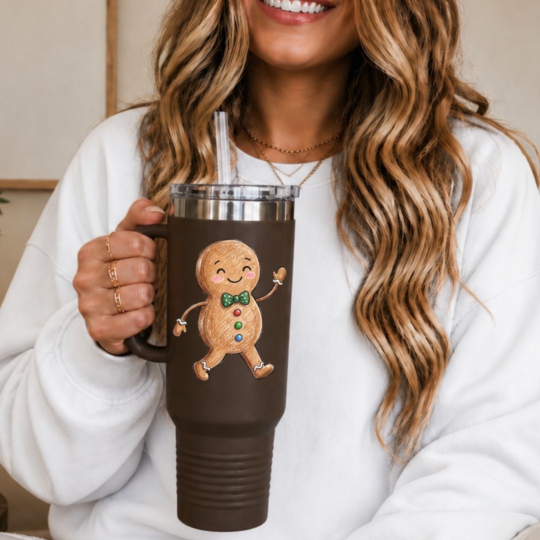 A woman in a white sweatshirt holds a brown insulated tumbler with a clear straw. The tumbler has a printed gingerbread man design with a green bow tie and colorful buttons.
