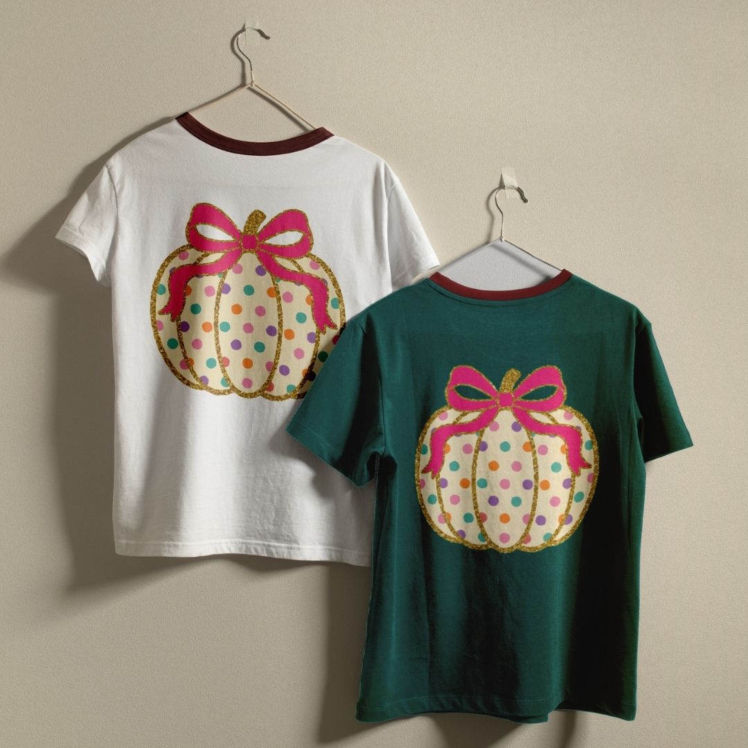 Two short-sleeve ringer t-shirts hanging on hangers against a plain beige wall. The left shirt is white with a dark red collar and features a large pumpkin graphic on the back with a pink bow and multicolored polka dots.