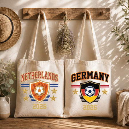Two cream canvas tote bags hanging from a wooden coat rack, one printed with a vintage orange Netherlands soccer crest and the other with a Germany soccer graphic.