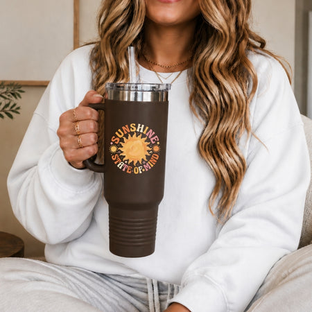 Person holding a brown mug with a sun design and text, wearing a white sweatshirt.