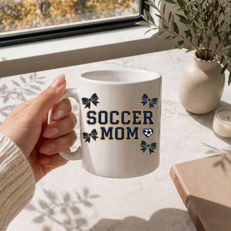 Hand holding a white mug with 'Soccer Mom' text and bow designs on a light surface with a window and plant in the background.