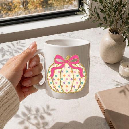 A hand holds a white ceramic mug featuring a pumpkin graphic with pastel polka dots, a pink bow, and gold glitter outlines, set on a light marble countertop near a window.