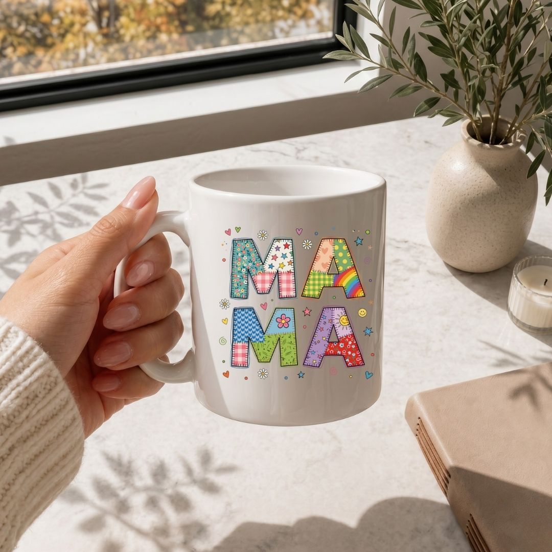 Hand holding a mug with 'MAMA' in colorful letters on a table with a window and plant in the background.