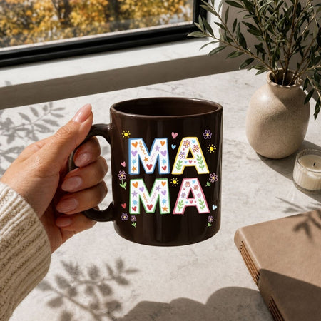 Hand holding a black mug with 'MAMA' in colorful letters on a table with a notebook and plant.