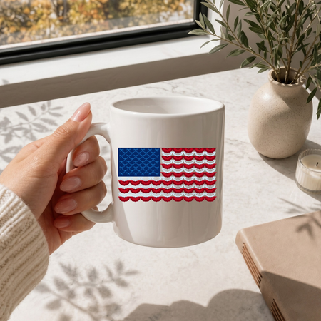 Hand holding a white mug with an American flag design on a light surface with a window and plant in the background.