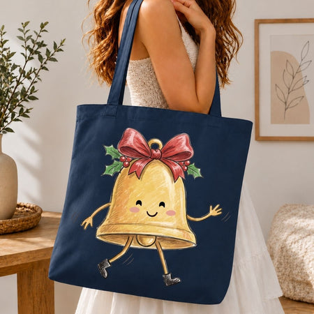 A navy blue canvas tote bag with a cute cartoon Christmas bell graphic, carried by a woman in a white dress in a cozy indoor setting.
