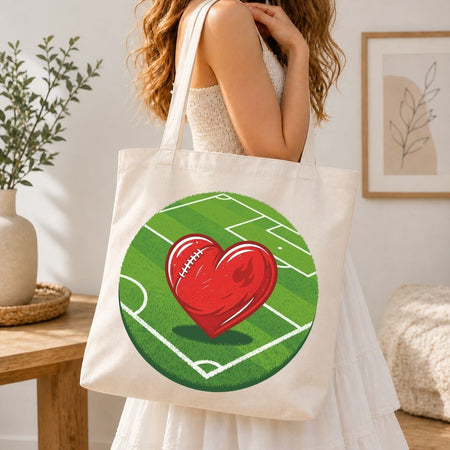 Person holding a tote bag with a heart and football design on a grass field.