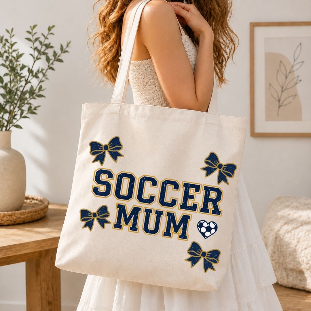 Person holding a tote bag with 'Soccer Mum' text and decorative elements in a home setting.