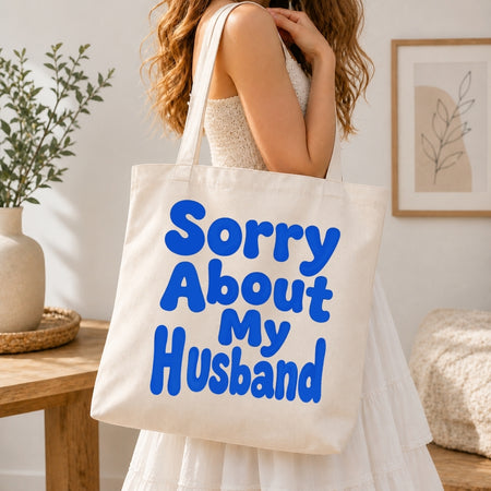 Person holding a tote bag with 'Sorry About My Husband' text in a home setting