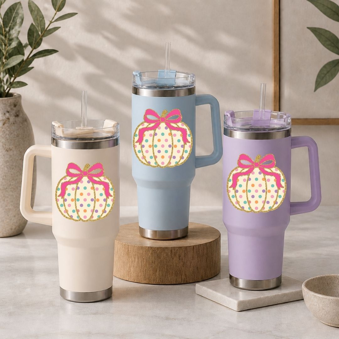 Three insulated travel tumblers with lids and straws are displayed on a tabletop. Each tumbler has a large pumpkin graphic with a pink bow and multicolored polka dots.