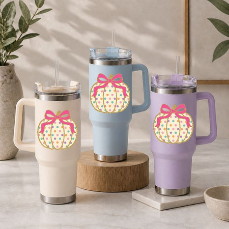Three insulated travel tumblers with lids and straws are displayed on a tabletop. Each tumbler has a large pumpkin graphic with a pink bow and multicolored polka dots.