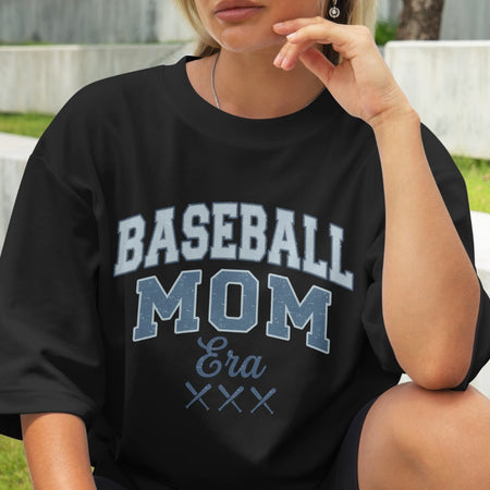 Person wearing a black t-shirt with 'BASEBALL MOM Era' text, sitting outdoors.