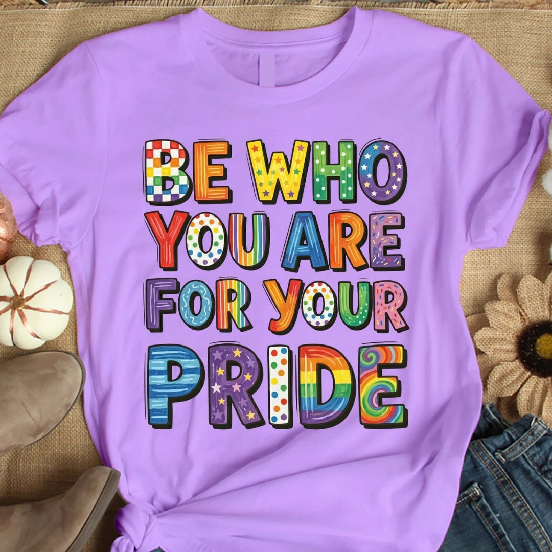 Purple t-shirt with colorful text 'Be who you are for your pride' on a textured surface.