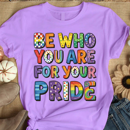Purple t-shirt with colorful text 'Be who you are for your pride' on a textured surface.