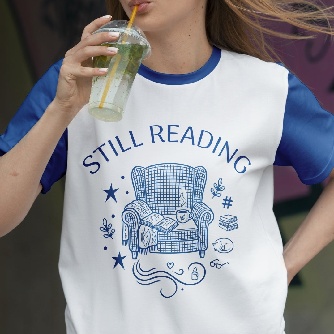 Person wearing a t-shirt with 'STILL READING' and armchair graphic, holding a drink.