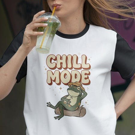 Person wearing a white t-shirt with 'Chill Mode' and a frog graphic, holding a drink.