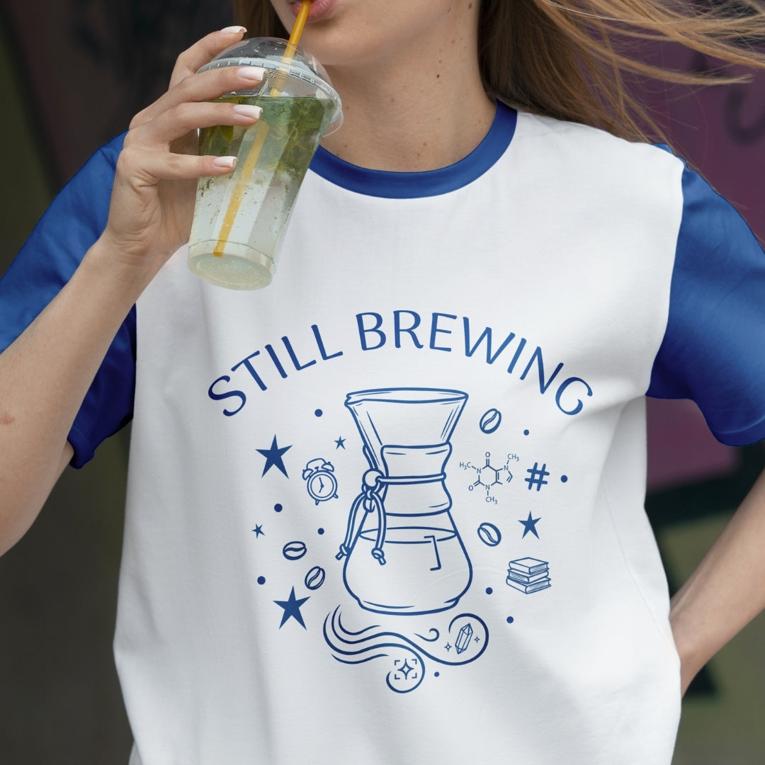 Person wearing a white t-shirt with blue sleeves and 'STILL BREWING' design, holding a drink.