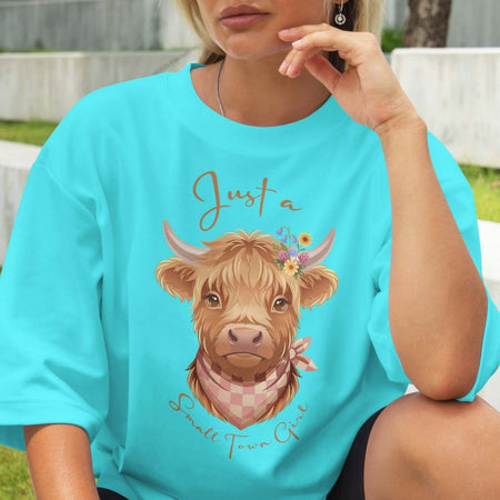 Person wearing a turquoise t-shirt with a highland cow graphic and text outdoors.