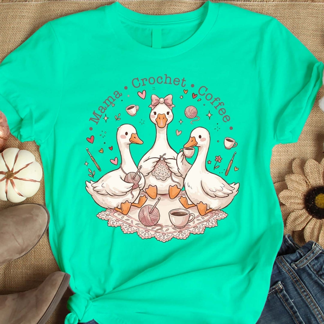 Turquoise t-shirt with a graphic of three ducks and text 'Mama Crochet Coffee' on a wooden surface.