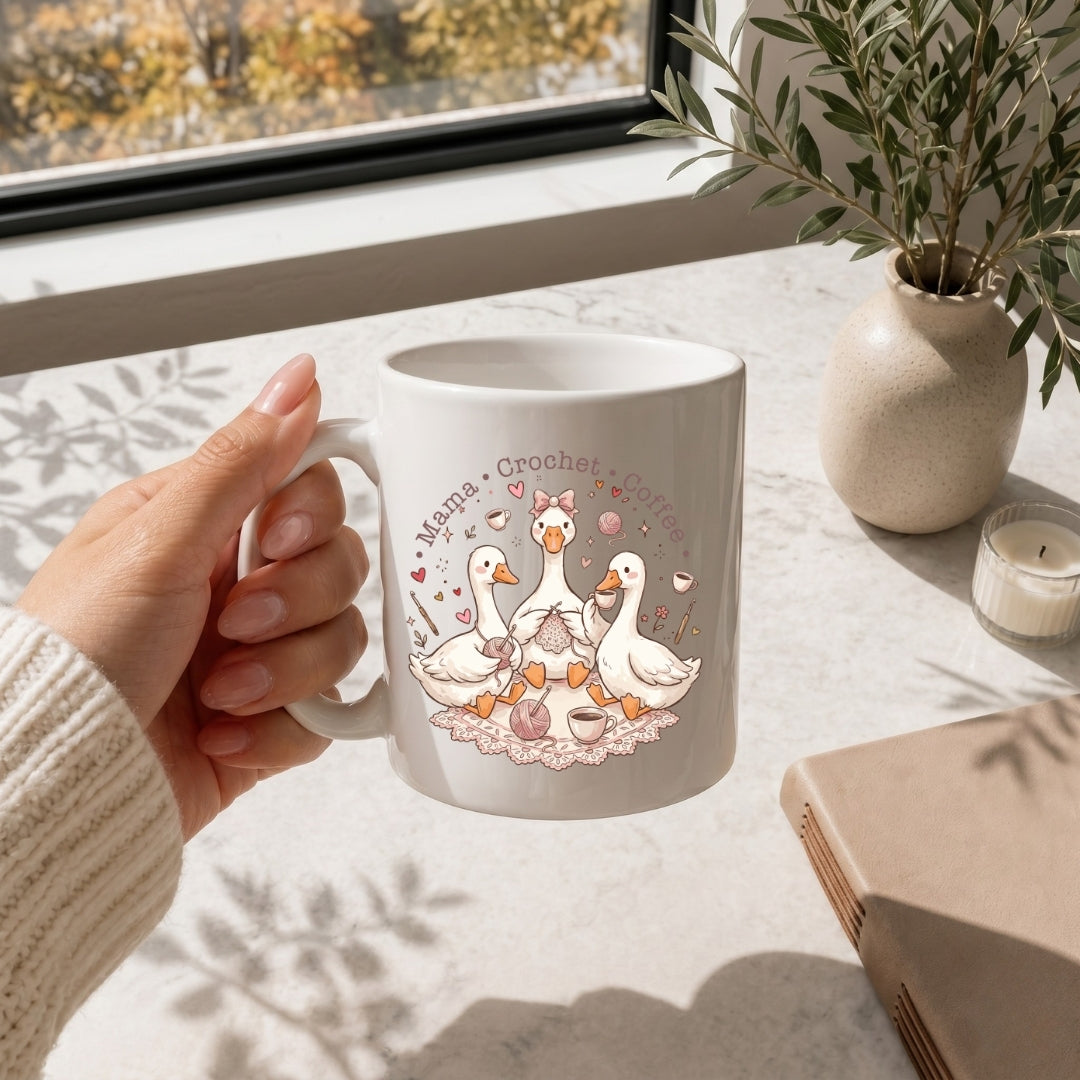 Person holding a mug with a 'Mama Crochet' design featuring ducks, sitting on a table with a window and plant in the background.