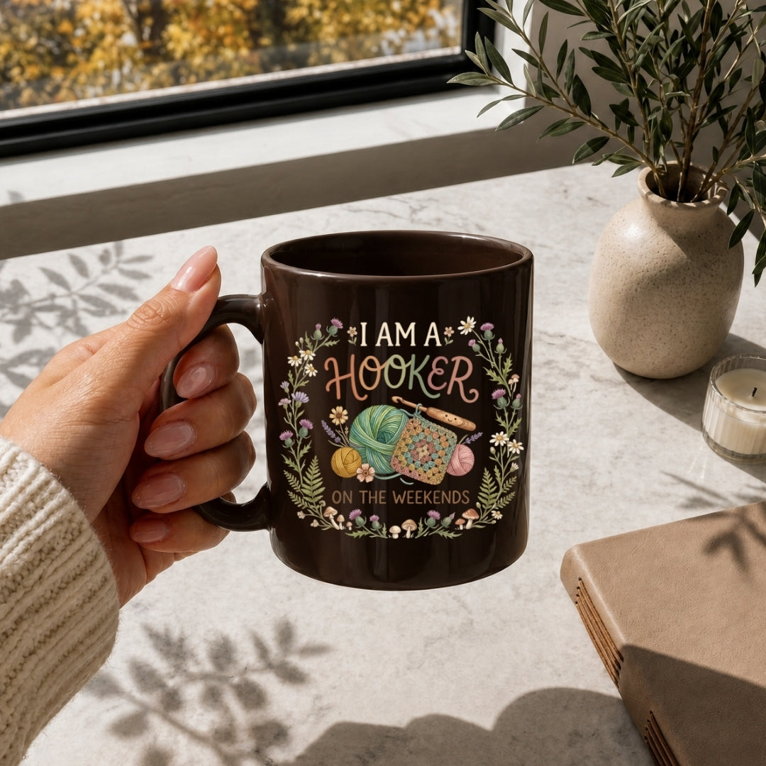 Person holding a mug with a crochet-themed design and text, placed on a table with a plant and notebook.