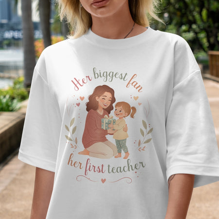 Person wearing a white t-shirt with a graphic and text 'Her biggest fan, her first teacher' outdoors.