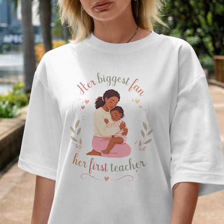 Person wearing a white t-shirt with a graphic of a woman holding a child and text 'Her biggest fan, her first teacher'.