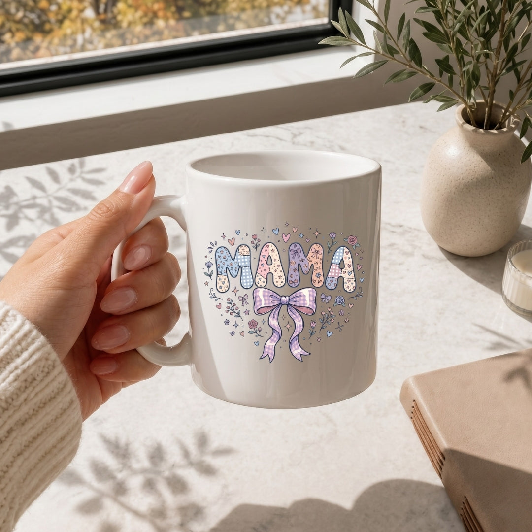 Hand holding a white mug with 'MAMA' and a bow design, placed on a light surface with a window and plant in the background.