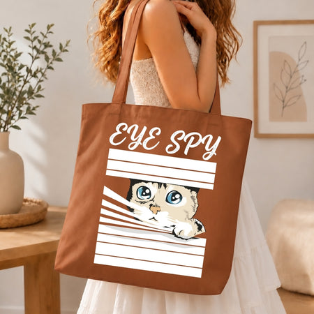 Brown tote bag with a cat design and 'Eye Spy' text held by a person in a home setting.
