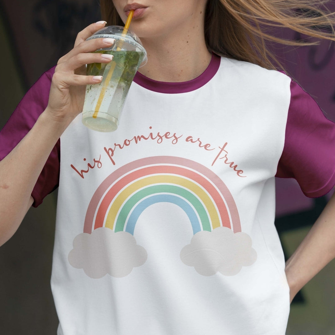 Person wearing a t-shirt with a rainbow and cloud design, holding a drink.