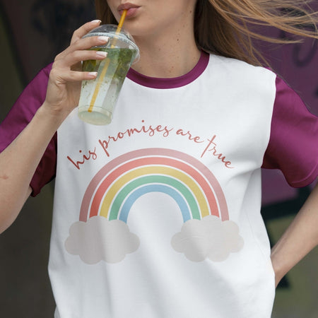 Person wearing a t-shirt with a rainbow and cloud design, holding a drink.