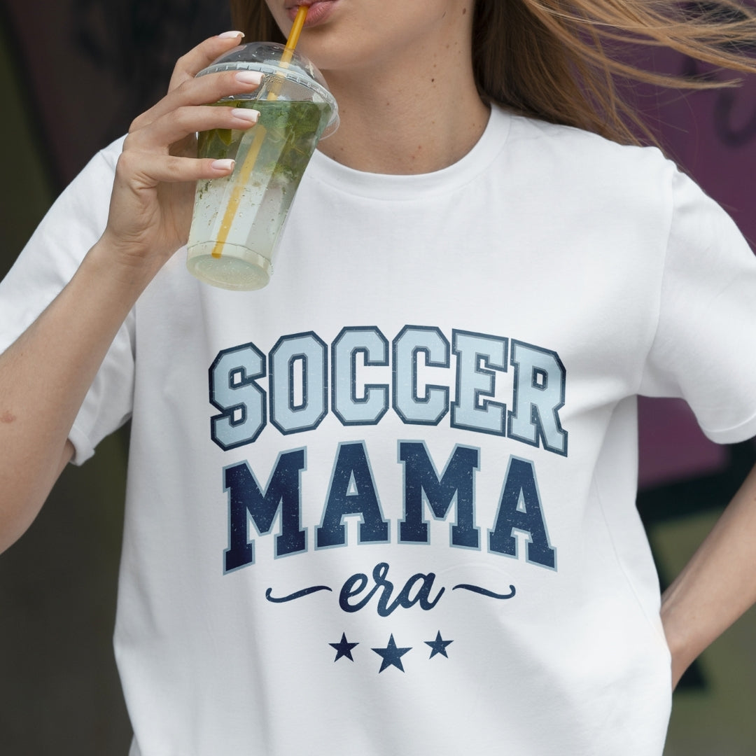 Person wearing a white t-shirt with 'Soccer Mama era' text, holding a drink.