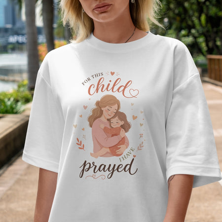White t-shirt with a graphic of a woman holding a child and text 'For this child I have prayed' worn by a person outdoors.