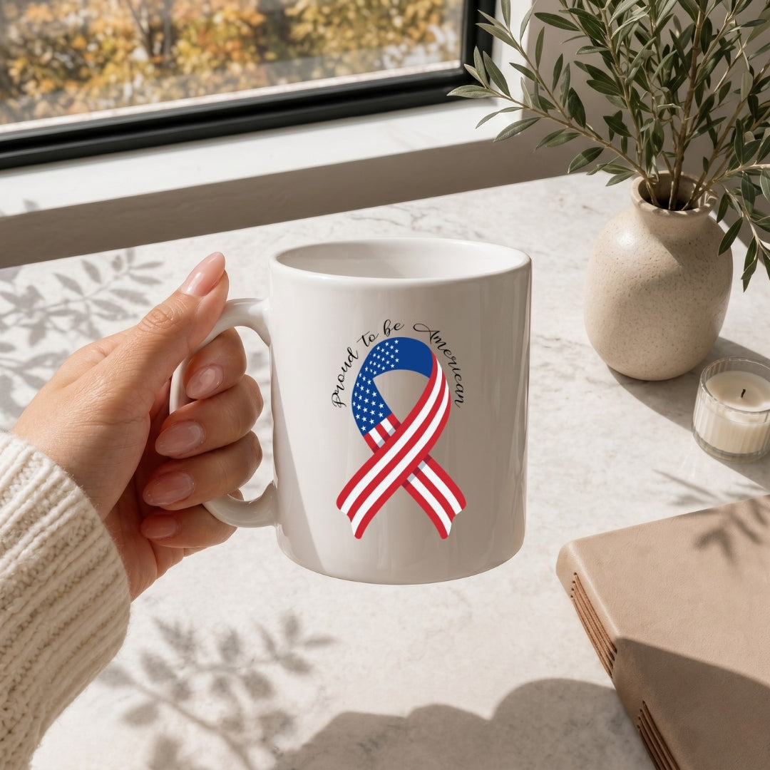 White mug with American flag ribbon design held by a hand on a table with a window and plant in the background.