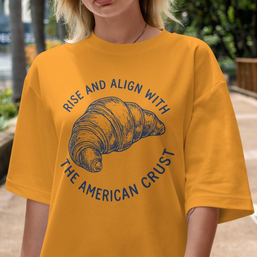 Person wearing a mustard yellow t-shirt with a croissant graphic and text outdoors.