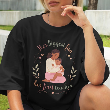 Person wearing a black t-shirt with a graphic of a woman holding a child and text 'Her biggest fan, her first teacher'.