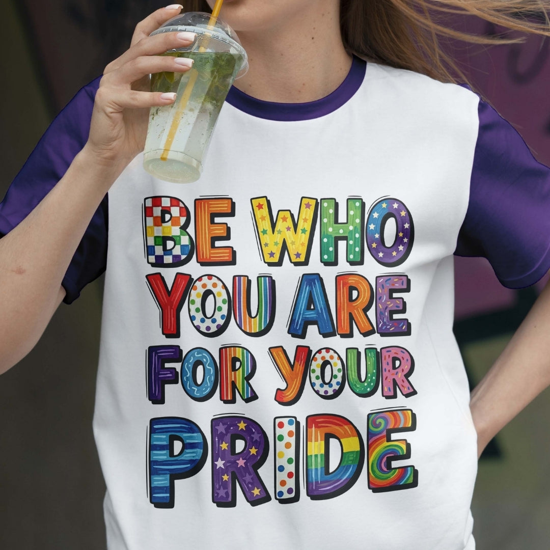 Person wearing a t-shirt with colorful text 'Be who you are for your pride' and holding a drink.
