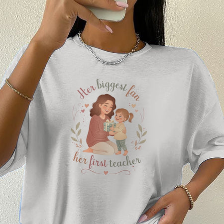 Person wearing a white t-shirt with a graphic and text 'Her biggest fan, her first teacher'.