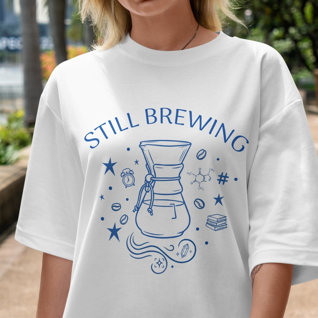 White t-shirt with 'STILL BREWING' design featuring a coffee filter and text, worn by a person outdoors.