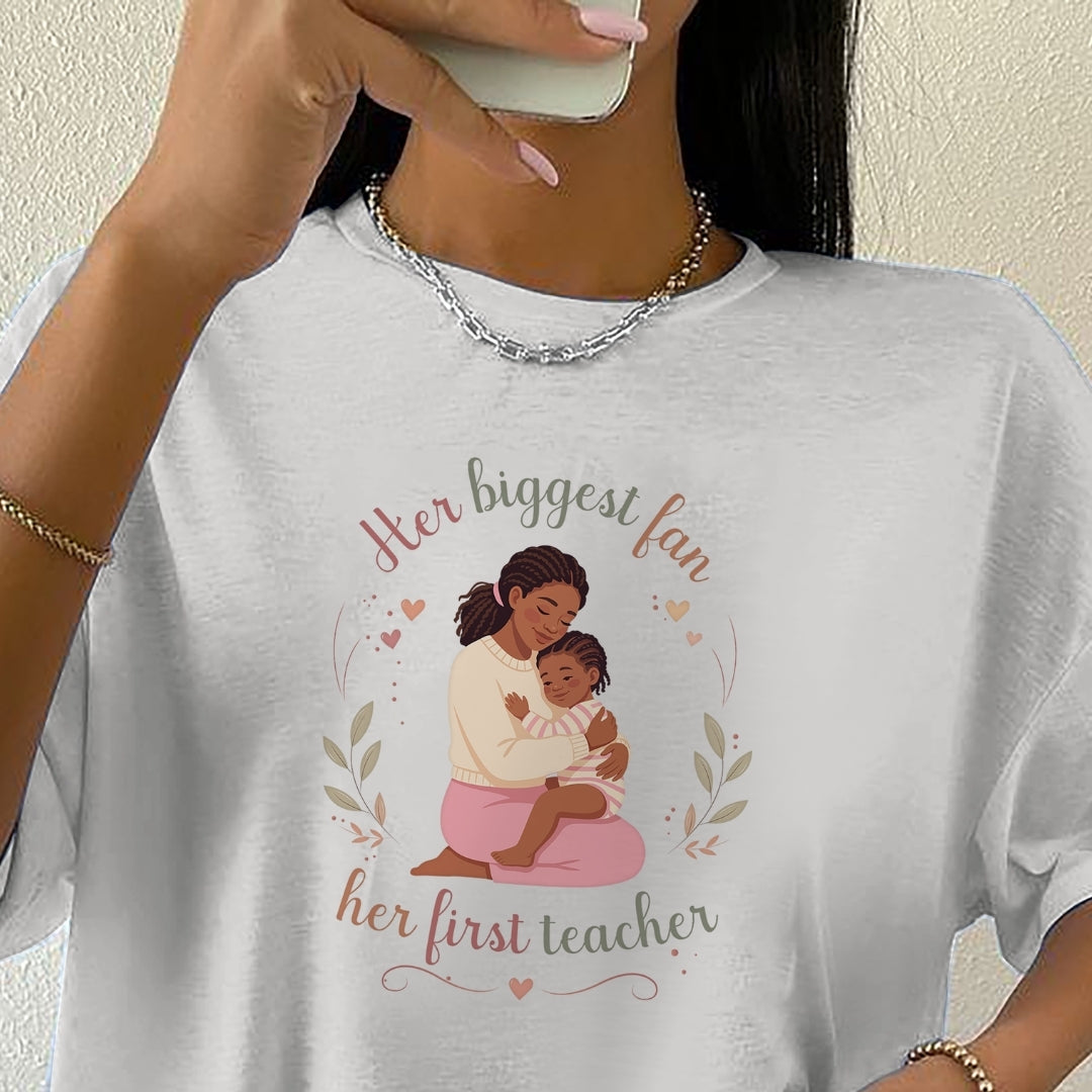 White t-shirt with a graphic of a woman holding a child and text 'Her biggest fan, her first teacher' on a plain background.