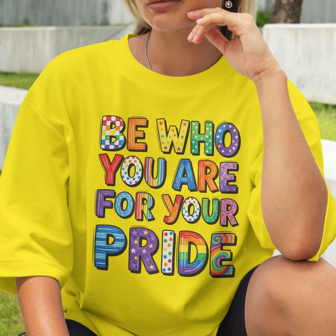 Person wearing a yellow t-shirt with colorful text 'BE WHO YOU ARE FOR YOUR PRIDE' outdoors.