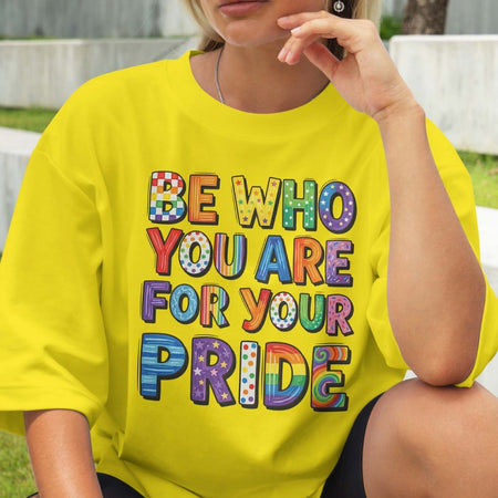 Person wearing a yellow t-shirt with colorful text 'BE WHO YOU ARE FOR YOUR PRIDE' outdoors.