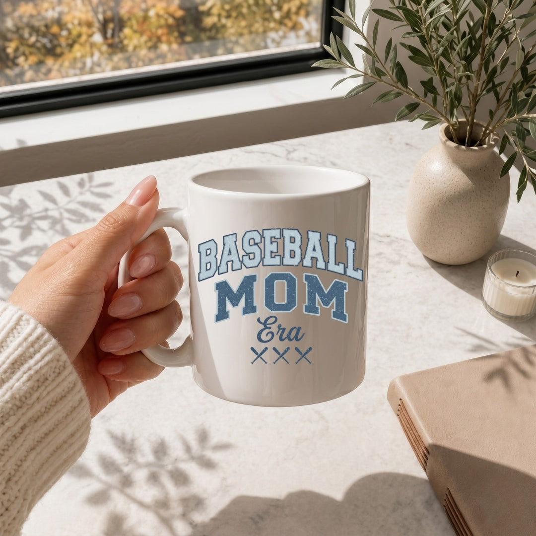 Person holding a 'Baseball Mom Era' mug on a table with a window and plant in the background