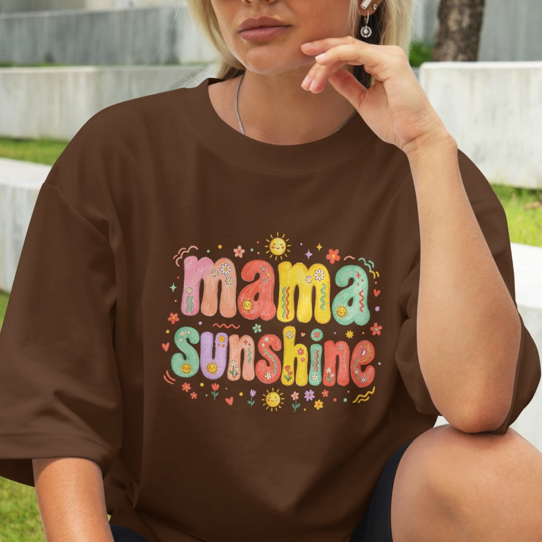 Close-up of a woman sitting outdoors wearing a chocolate brown oversized t-shirt printed with a vibrant, colorful 1970s style "Mama Sunshine" design.