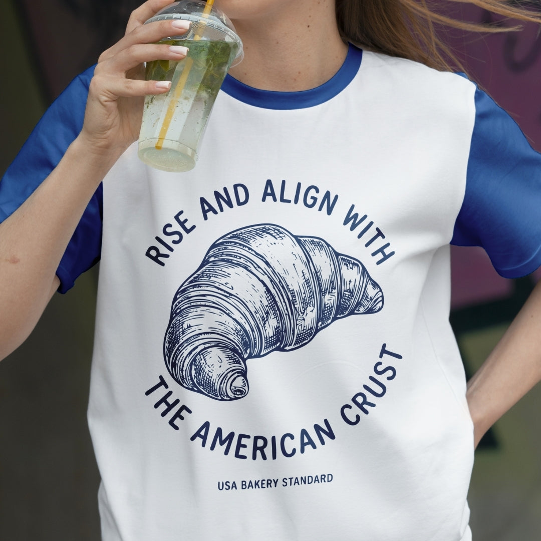 Person wearing a t-shirt with a croissant graphic and text, holding a drink.