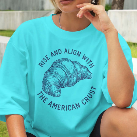 Person wearing a turquoise t-shirt with a croissant graphic and text outdoors.