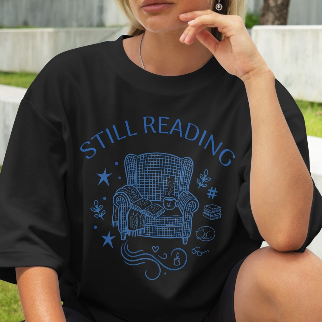 Person wearing a black t-shirt with 'STILL READING' and armchair graphic.